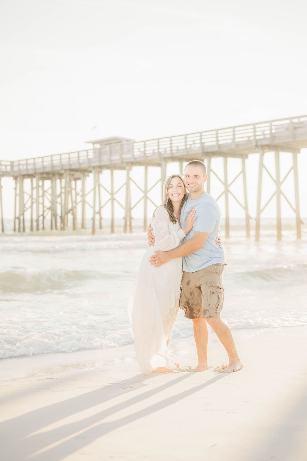 A loving couple embraces on a sunny beach near a wooden pier during golden hour.