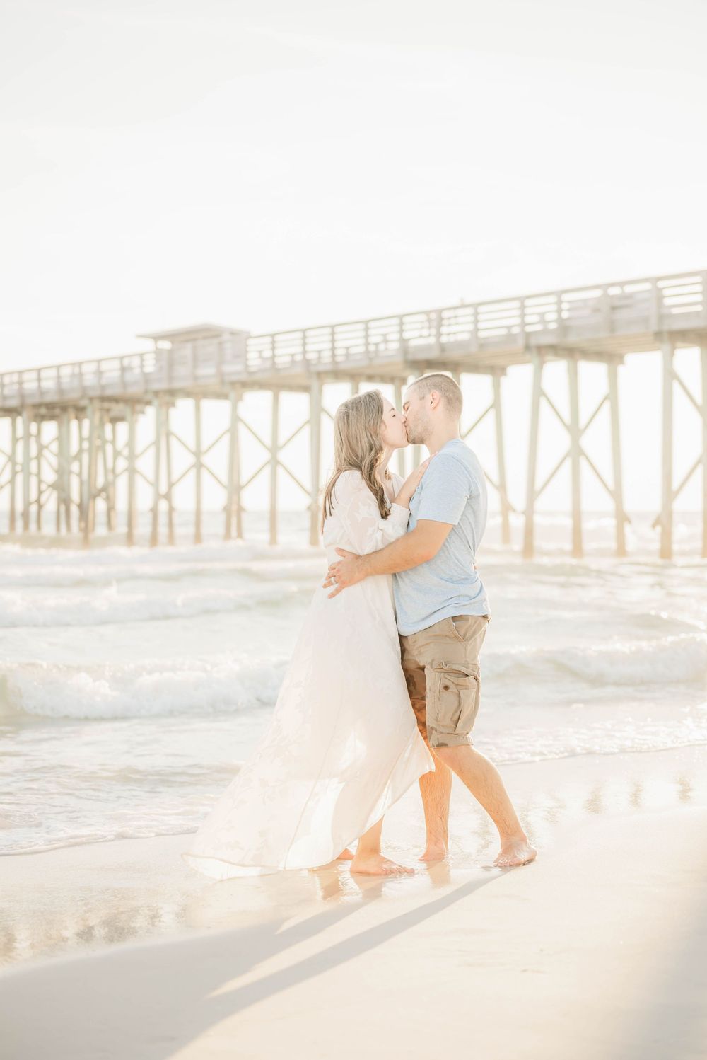 A couple embraces on a sunlit beach with a wooden pier in the background during a romantic photoshoot.