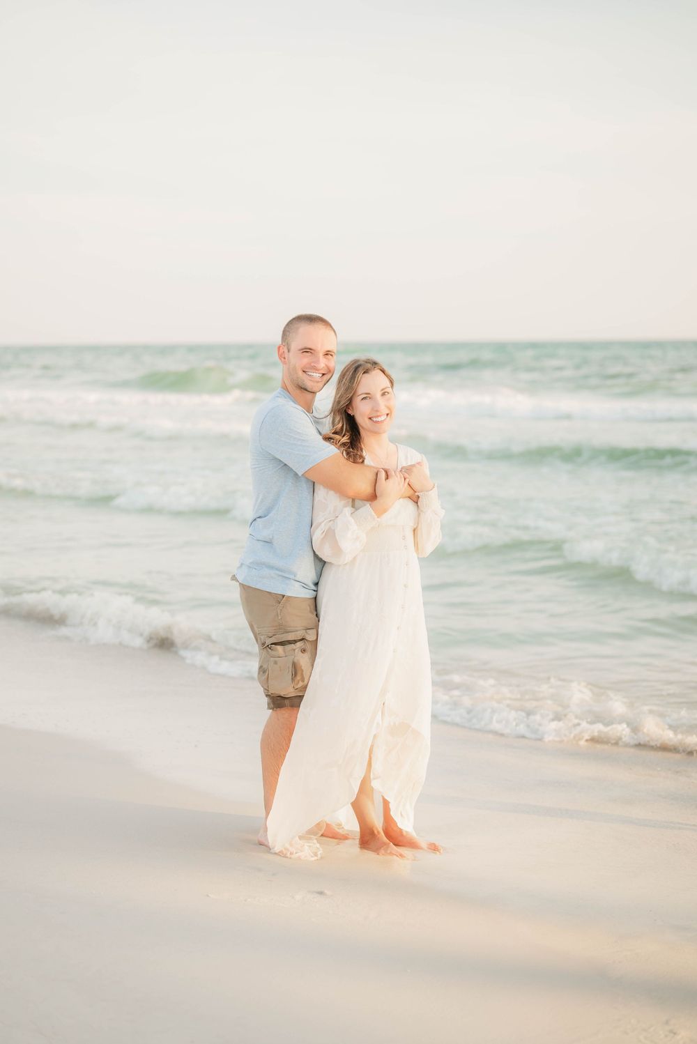 A couple embraces while standing barefoot on a peaceful beach at sunset with gentle waves rolling in.