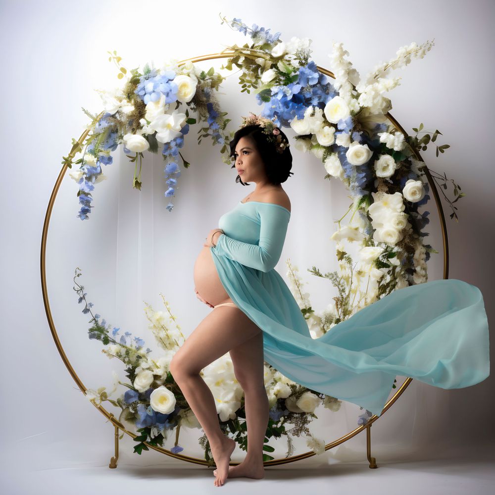 An expectant woman in a flowing turquoise dress poses by a round floral arch with white and blue blooms. Professional newborn photographer,
