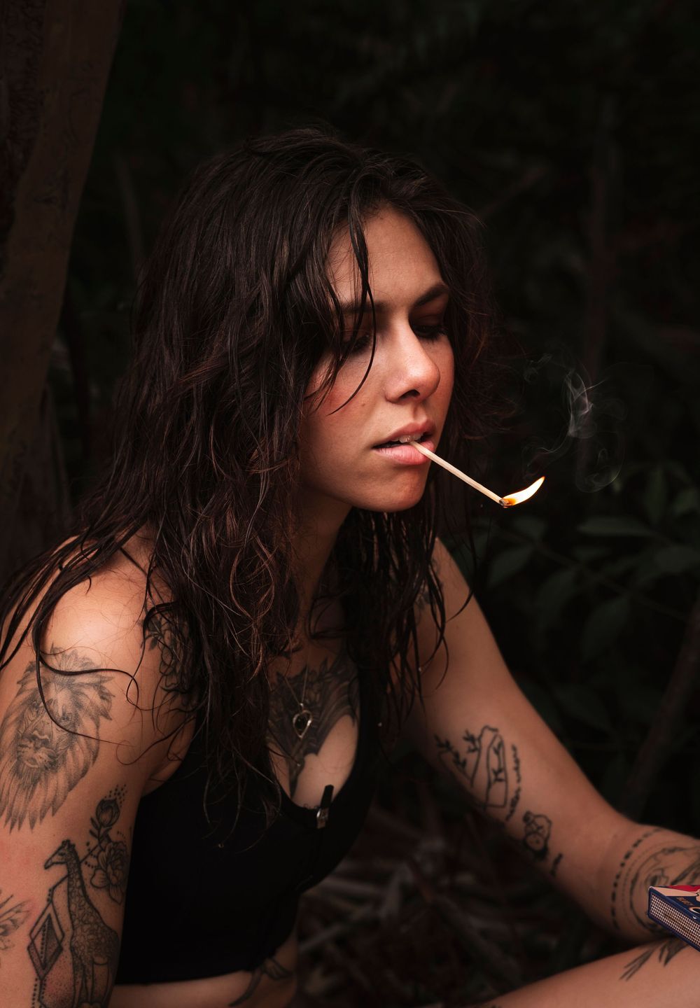 A person with dark hair and tattoos in a dark forest setting lights a match in a moody portrait.