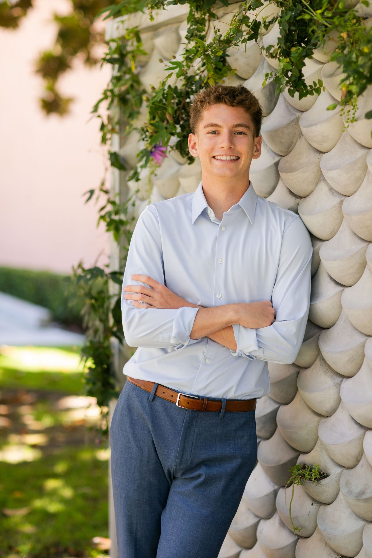 Professional portrait of someone in a light blue button-down shirt and navy slacks posing against a textured wall with greenery.