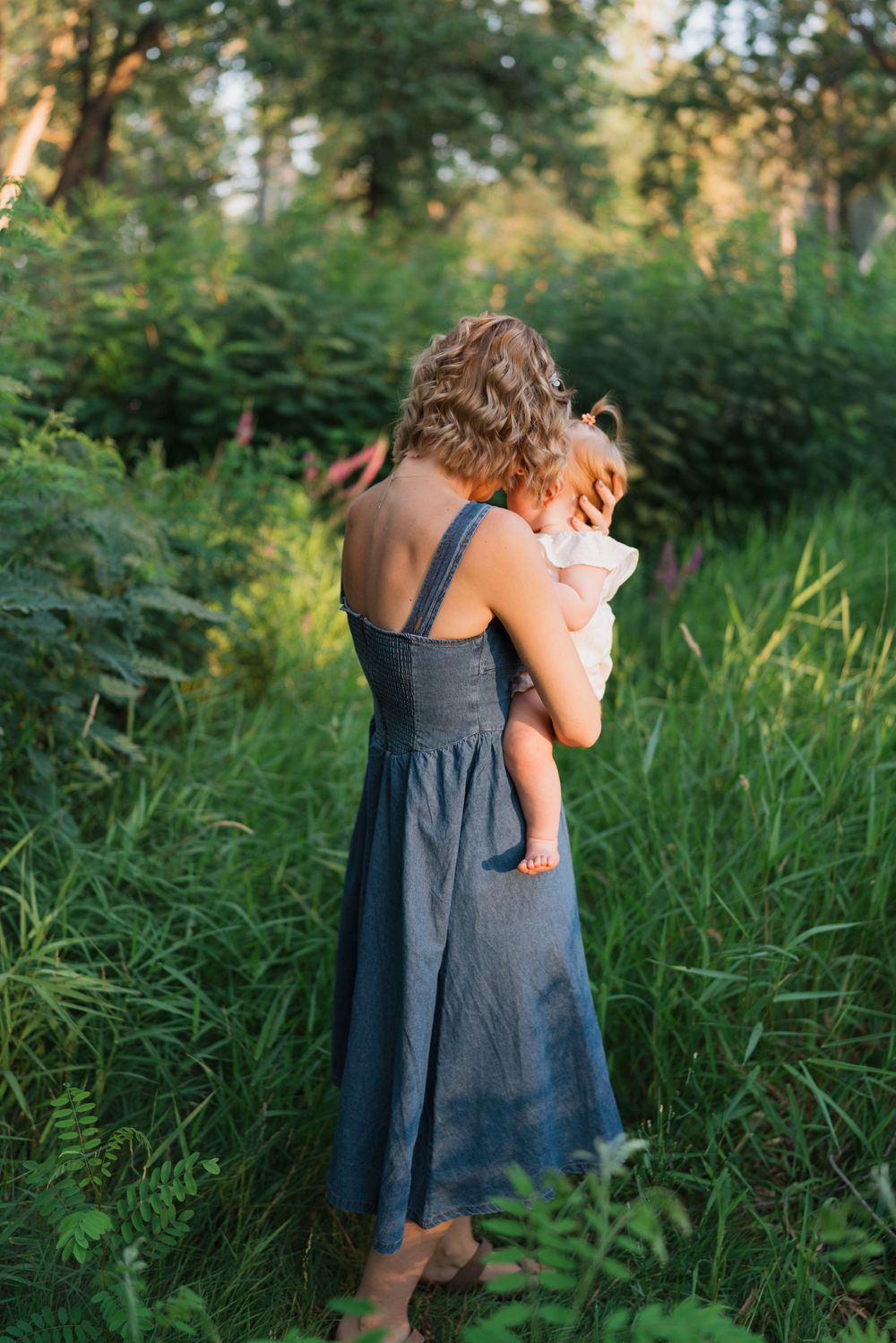 A mother in a blue dress holds her baby while standing in a lush green forest at golden hour.