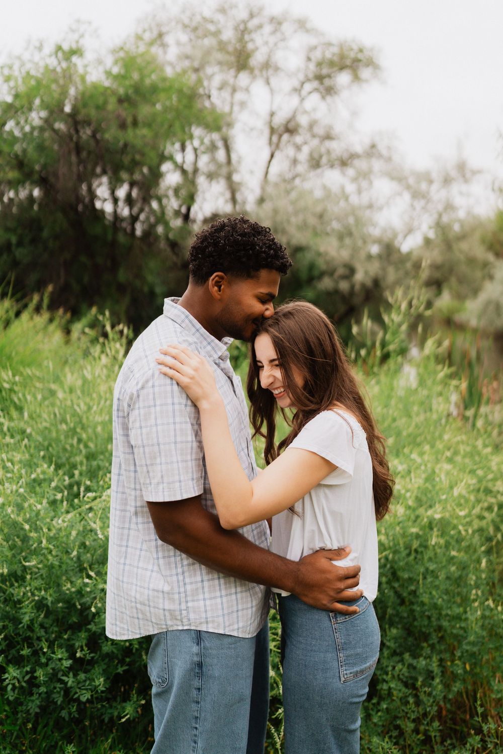 A loving interracial couple embraces and smiles together outdoors, surrounded by lush green foliage.