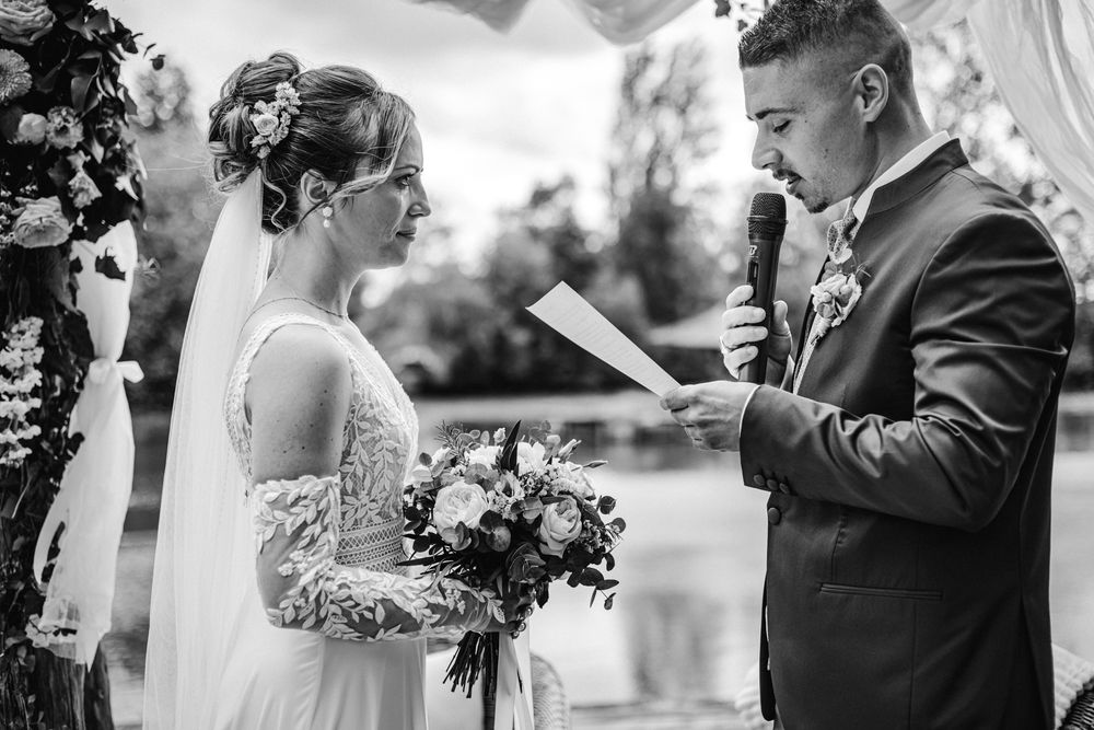 Groom reads wedding vows to bride holding bouquet at outdoor ceremony in black and white photo.