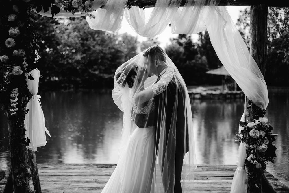 Black and white photo of bride and groom sharing first kiss under floral arch by lakeside at outdoor wedding.