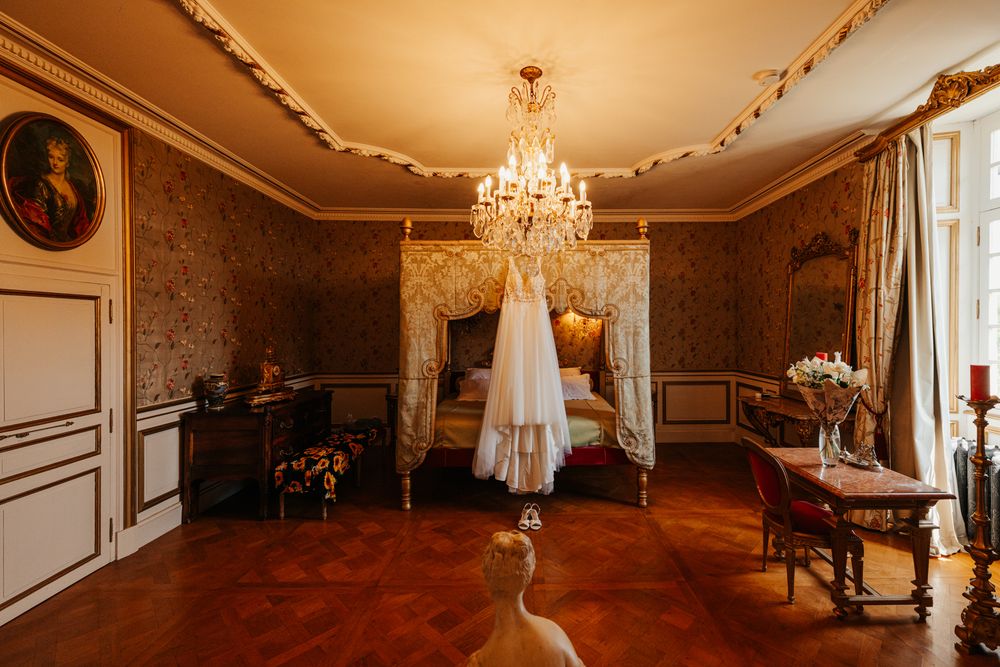 Elegant French château bridal suite with wedding dress hanging from ornate canopy bed under crystal chandelier.