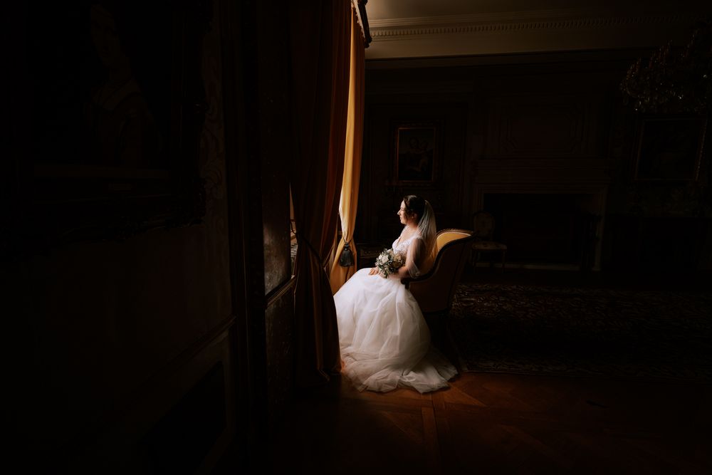 Bride in white gown sits by golden curtains in a dimly lit elegant room, holding a bouquet on her wedding day.