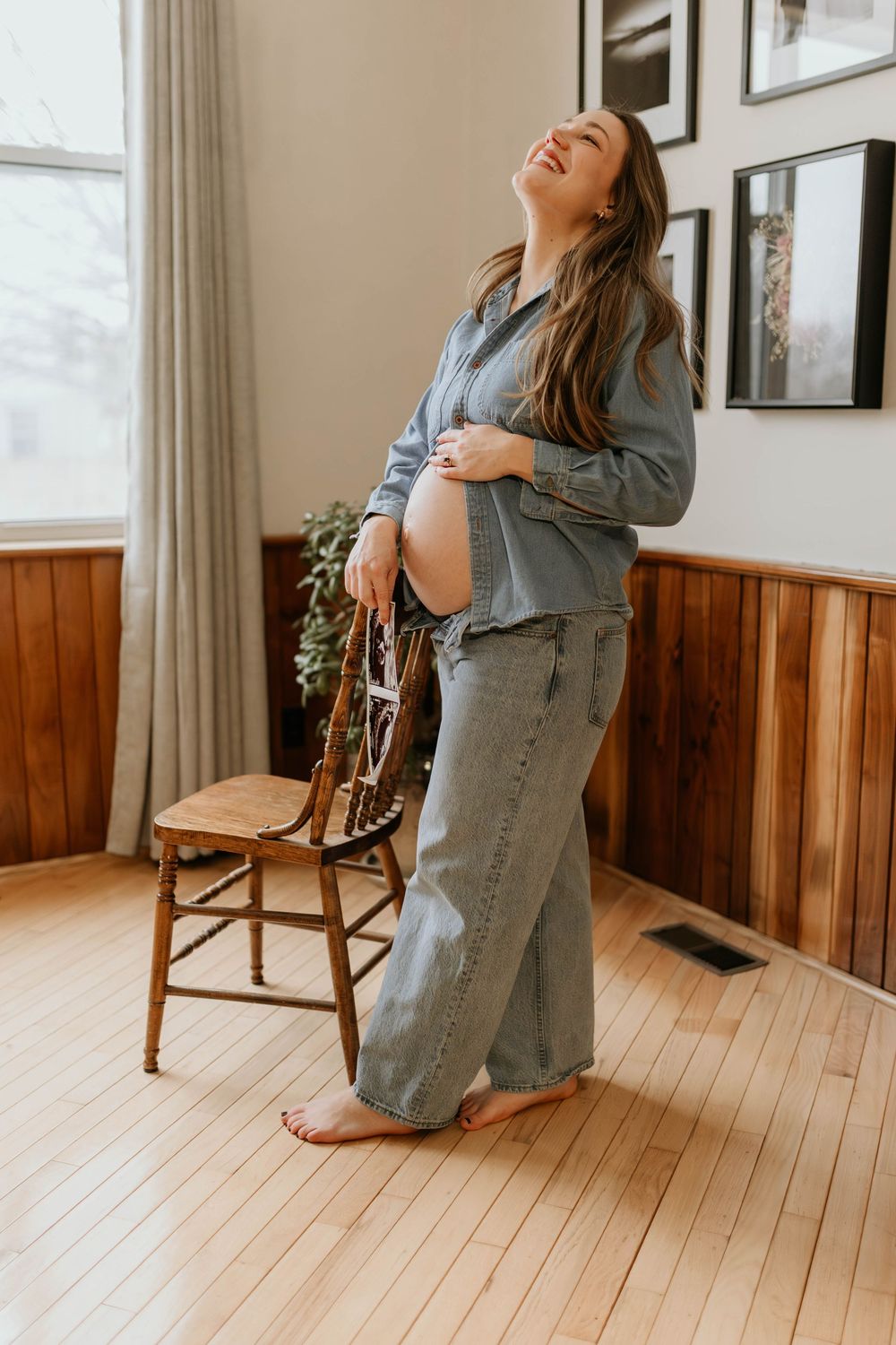 Expectant mother in denim outfit laughs joyfully while standing next to wooden chair in room with wainscoting.