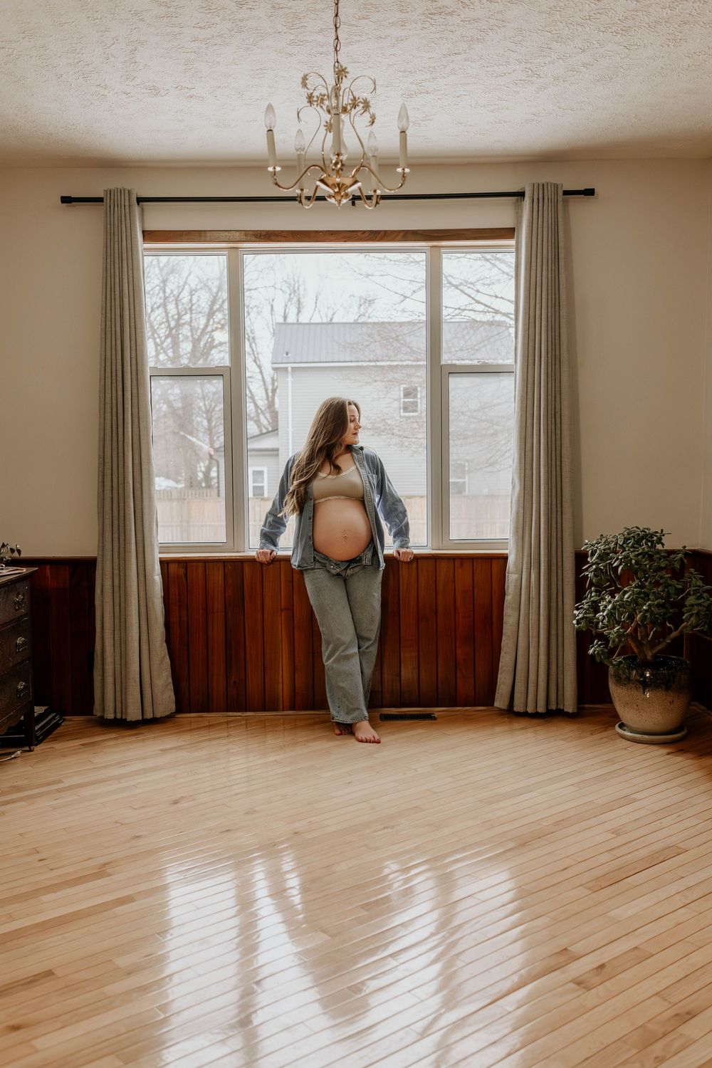 Pregnant woman in denim stands by window in room with hardwood floors and elegant chandelier.
