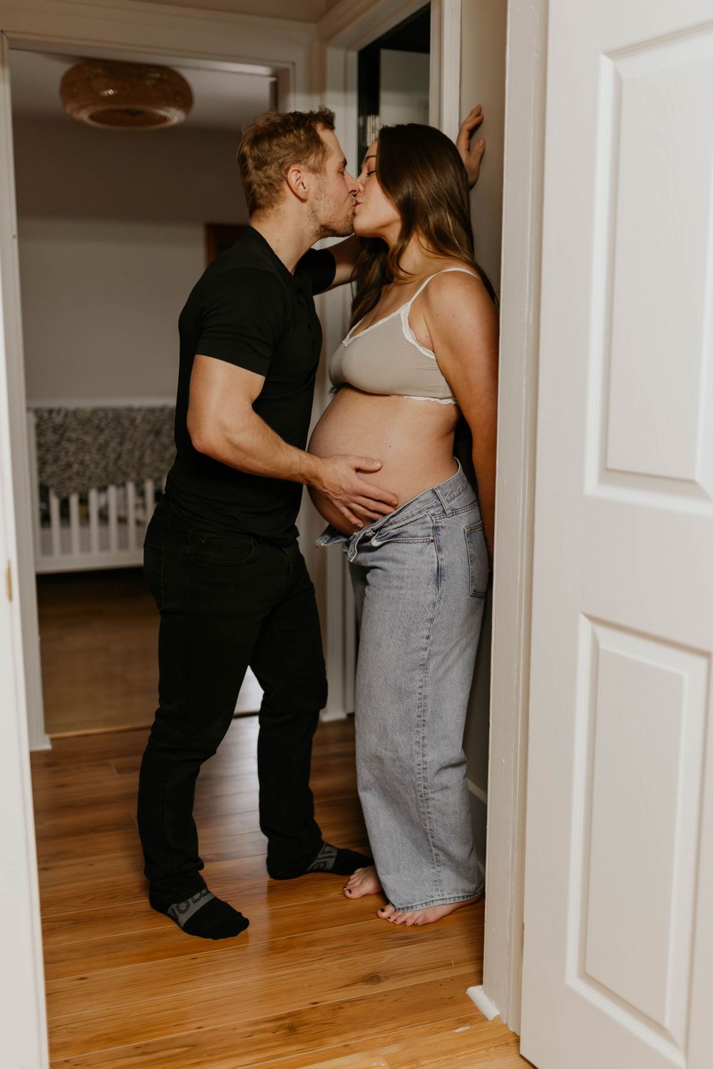 A couple embraces intimately in a hallway with hardwood floors and white walls, reflected in multiple mirror views.