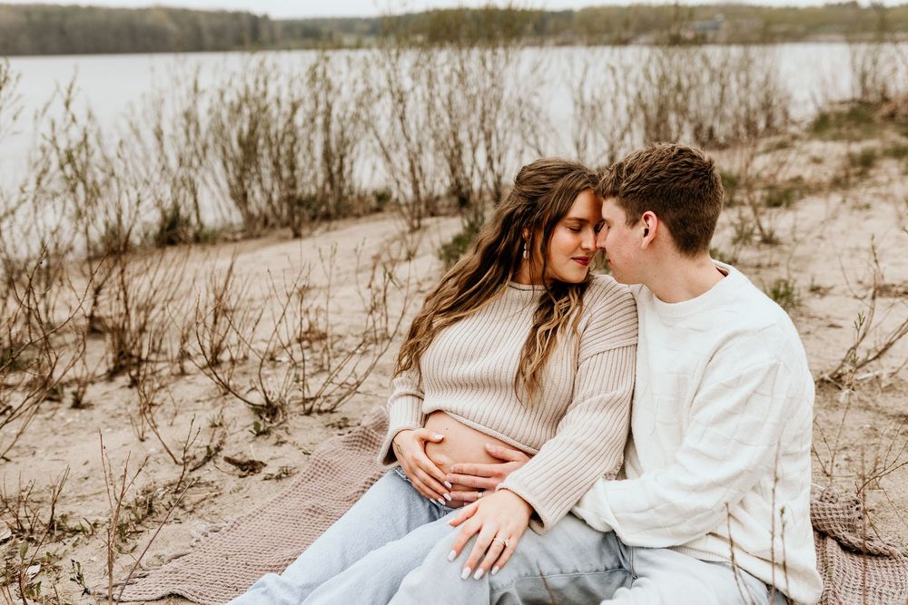 A romantic couple sits together on a sandy beach near tall grass and a lake on an overcast day.