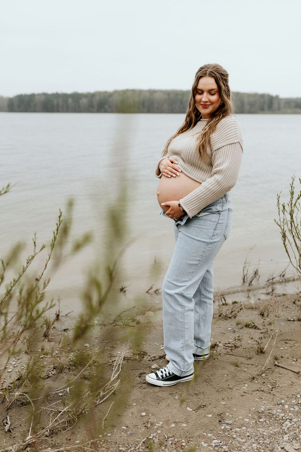 Someone wearing light beige sweater and blue jeans poses casually by a misty lakeside on an overcast day.