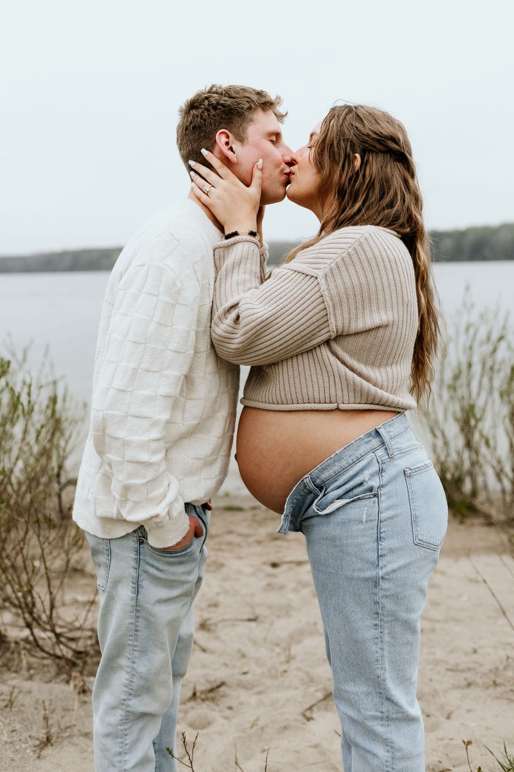 Couple in casual outfits sharing intimate moments on a beach with water and vegetation in background.