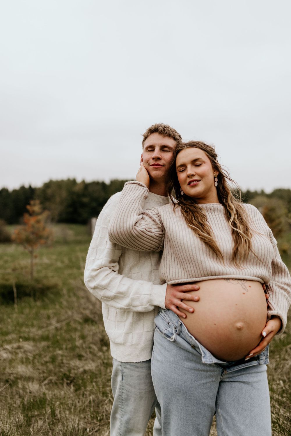 A maternity photo series showing a couple in cream sweaters embracing in a natural outdoor setting.