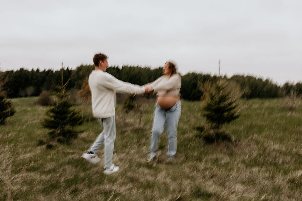Two people greet each other in an open field surrounded by evergreen trees on an overcast day.