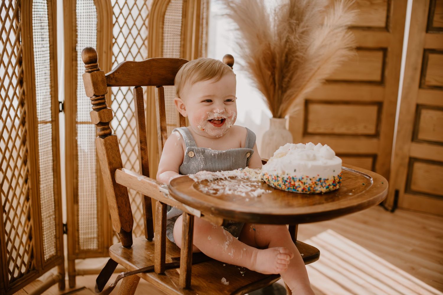 A toddler sitting in a wooden chair laughs joyfully next to a decorated cake and pampas grass decor.