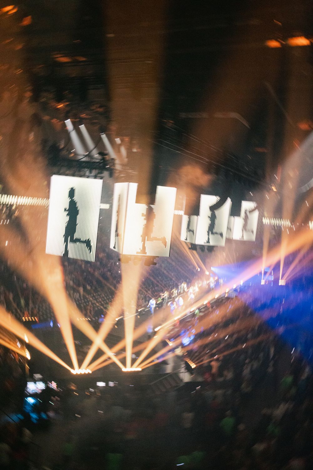 Aerial view of a concert arena with dramatic light beams, large screens, and a packed crowd bathed in golden light.
