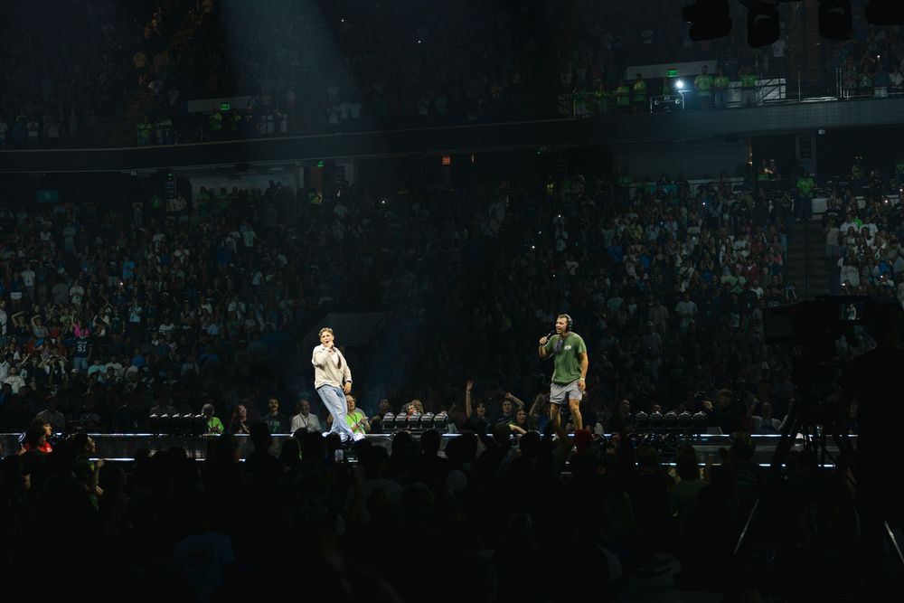Two performers on a large concert stage in a packed arena with crowds of fans in the background.