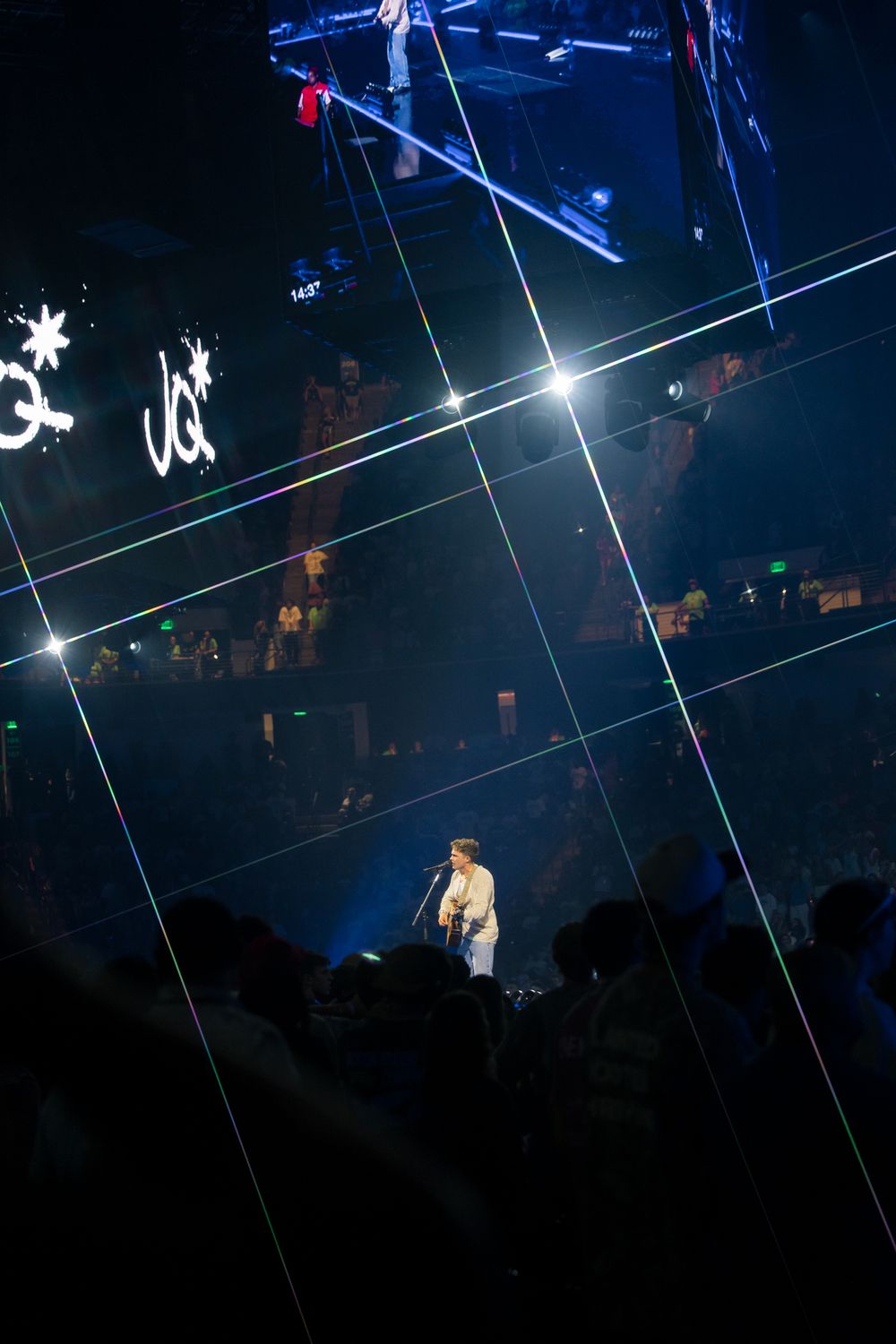 Performer on stage with dramatic laser light show and crowd silhouettes at a live concert venue.