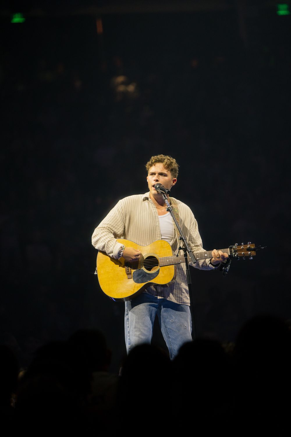 Male musician performing on stage, playing acoustic guitar and singing into microphone under spotlight.