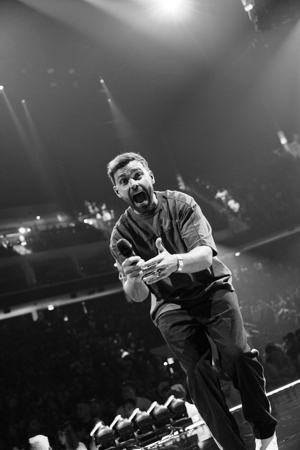 Black and white photo of an energetic performer on stage, leaning forward with open mouth, holding a microphone.