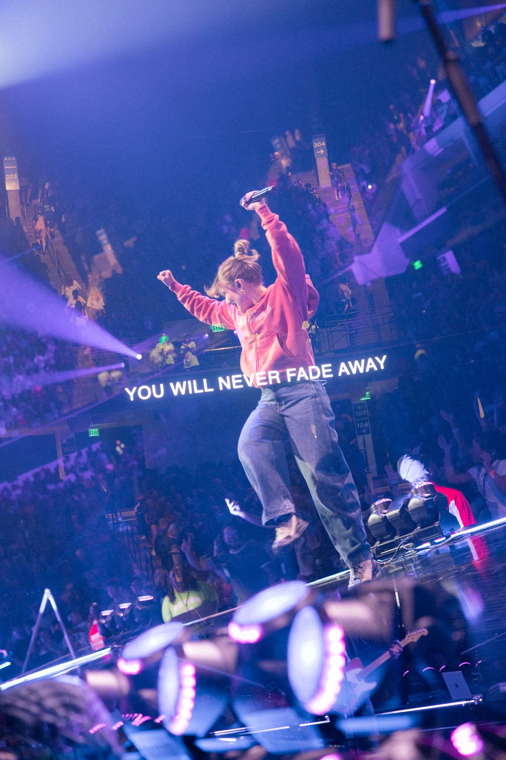 Performer on stage with arms raised, crowd in background, lit by purple lights, text reads YOU WILL NEVER FADE AWAY.