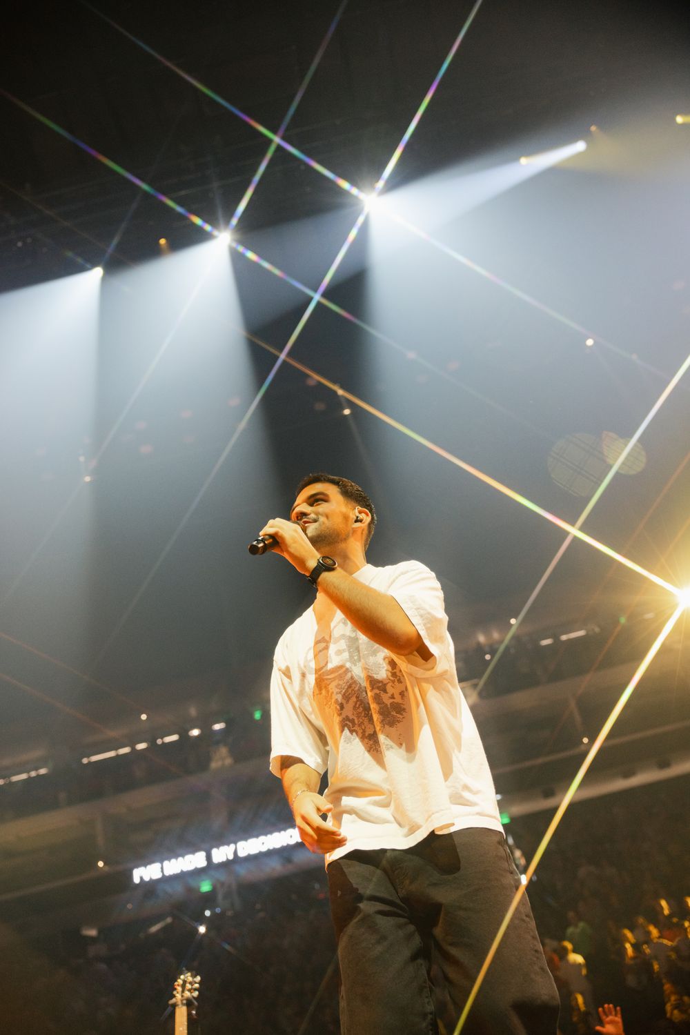 Performer in white t-shirt holds microphone on stage under colorful laser lights at a live concert.