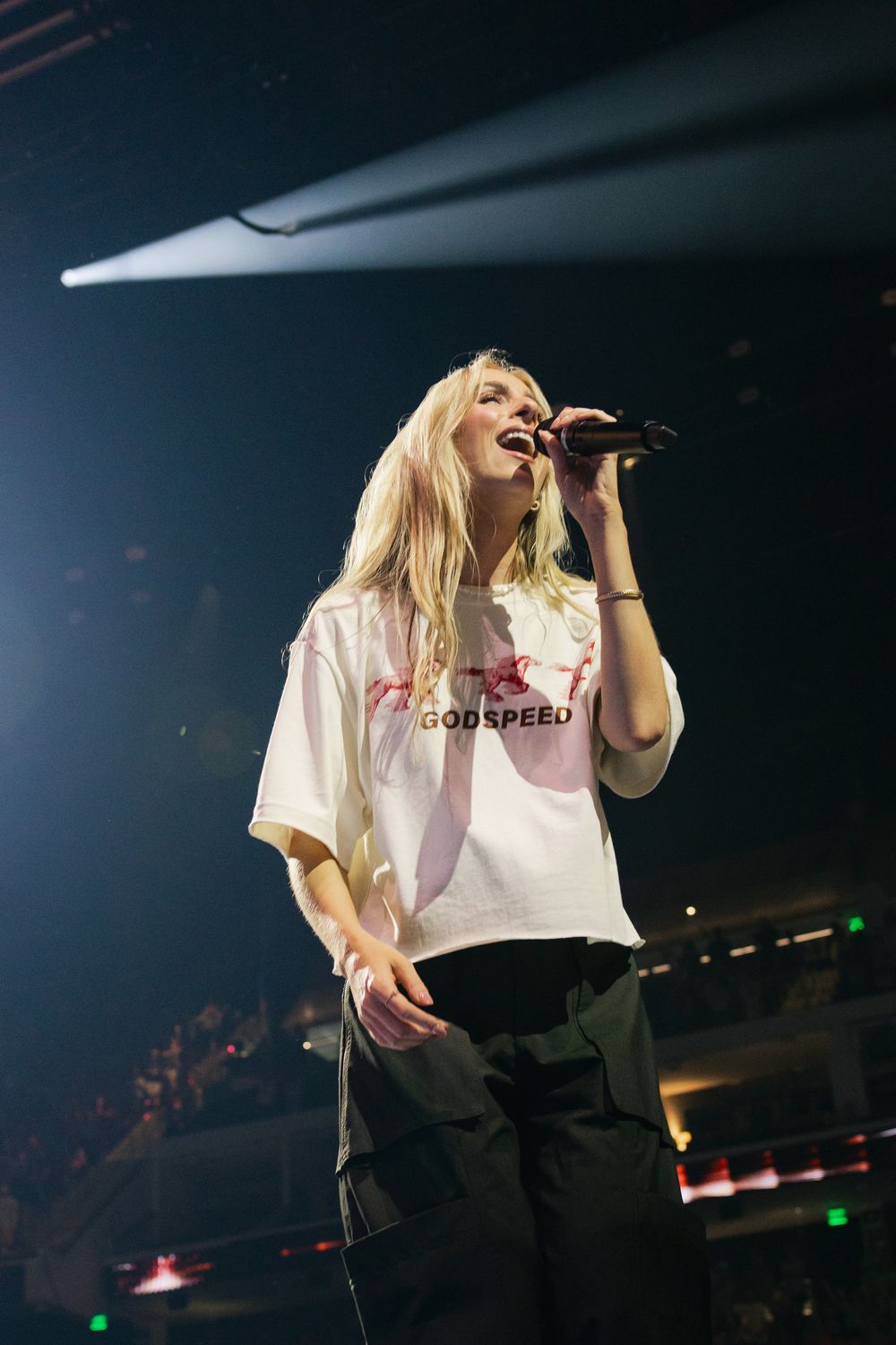 Blonde female singer performing on stage wearing a white Godspeed t-shirt, holding a microphone under dramatic stage lighting.