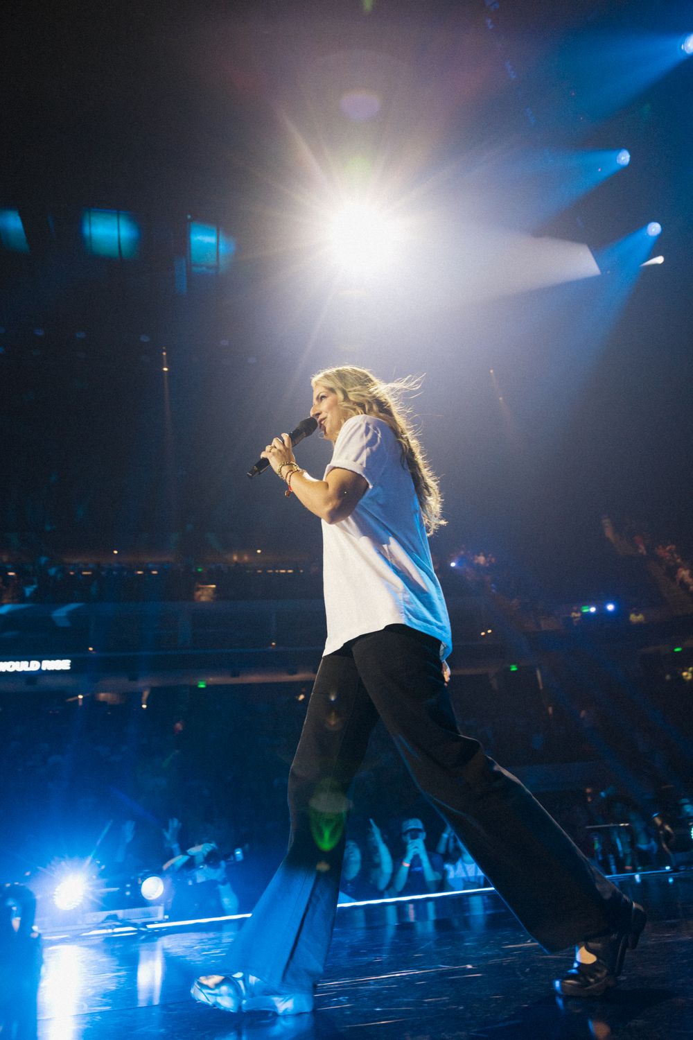 Female performer with long blonde hair singing on stage under bright spotlight in white top and black pants.