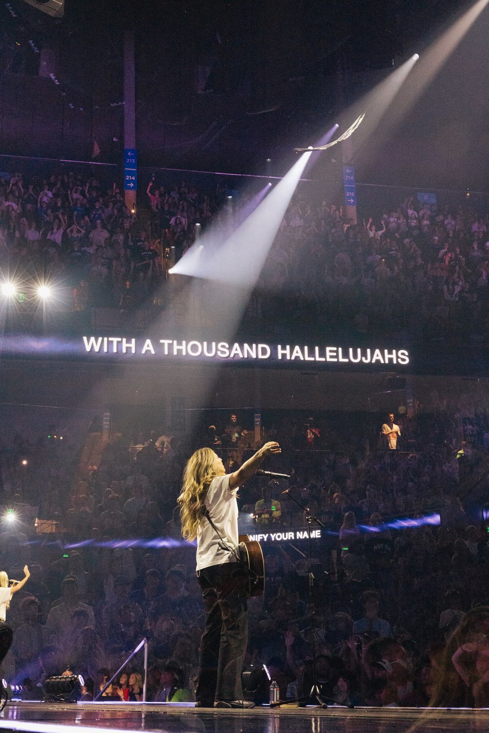 Worship leader on stage with spotlight, crowd in background, lyrics 'With A Thousand Hallelujahs' displayed on screen.
