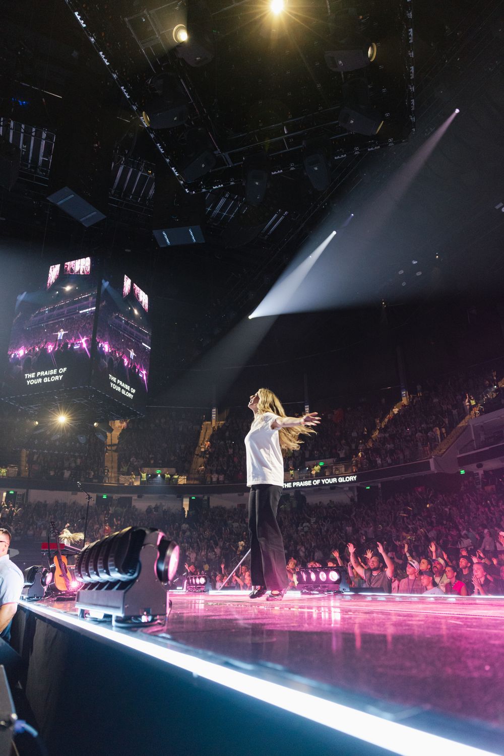 Performer on stage with arms outstretched, facing a large crowd at an indoor arena concert with pink lighting.