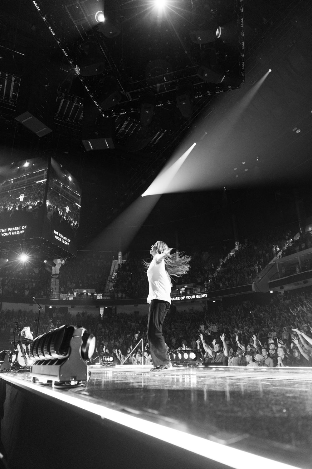 Black and white photo of a performer on stage with dramatic spotlight, large crowd visible in the background.