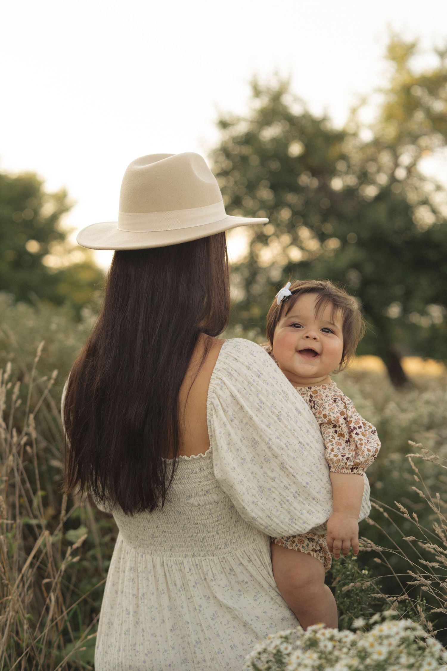 Behind view of mother in white dress and hat holding baby in sunlit field.