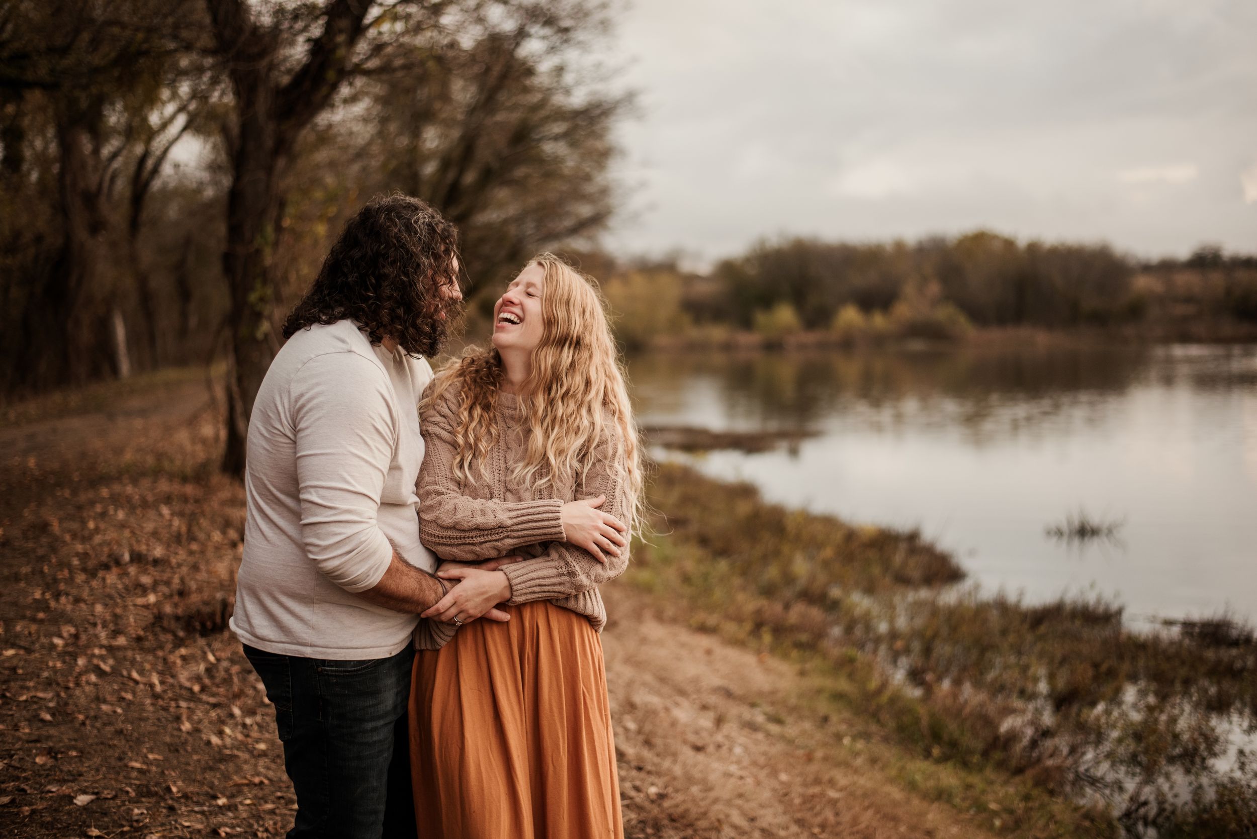 Couple in autumn attire embracing by a serene lake during golden hour photography session.