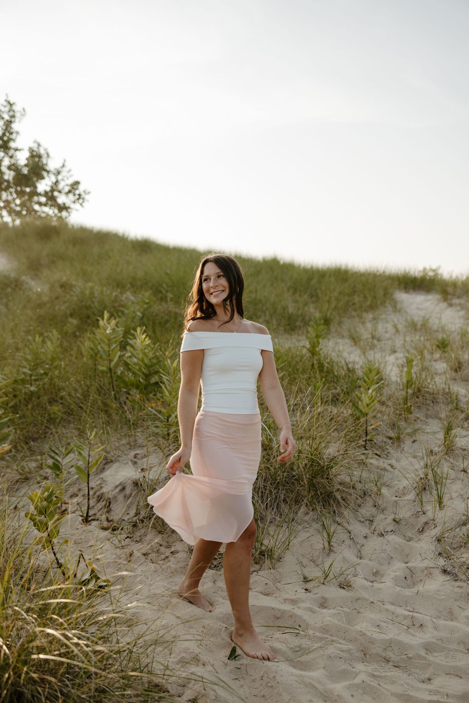 A woman in a white off-the-shoulder top and pink skirt walking on a sandy beach surrounded by vegetation. Fort Worth Texas