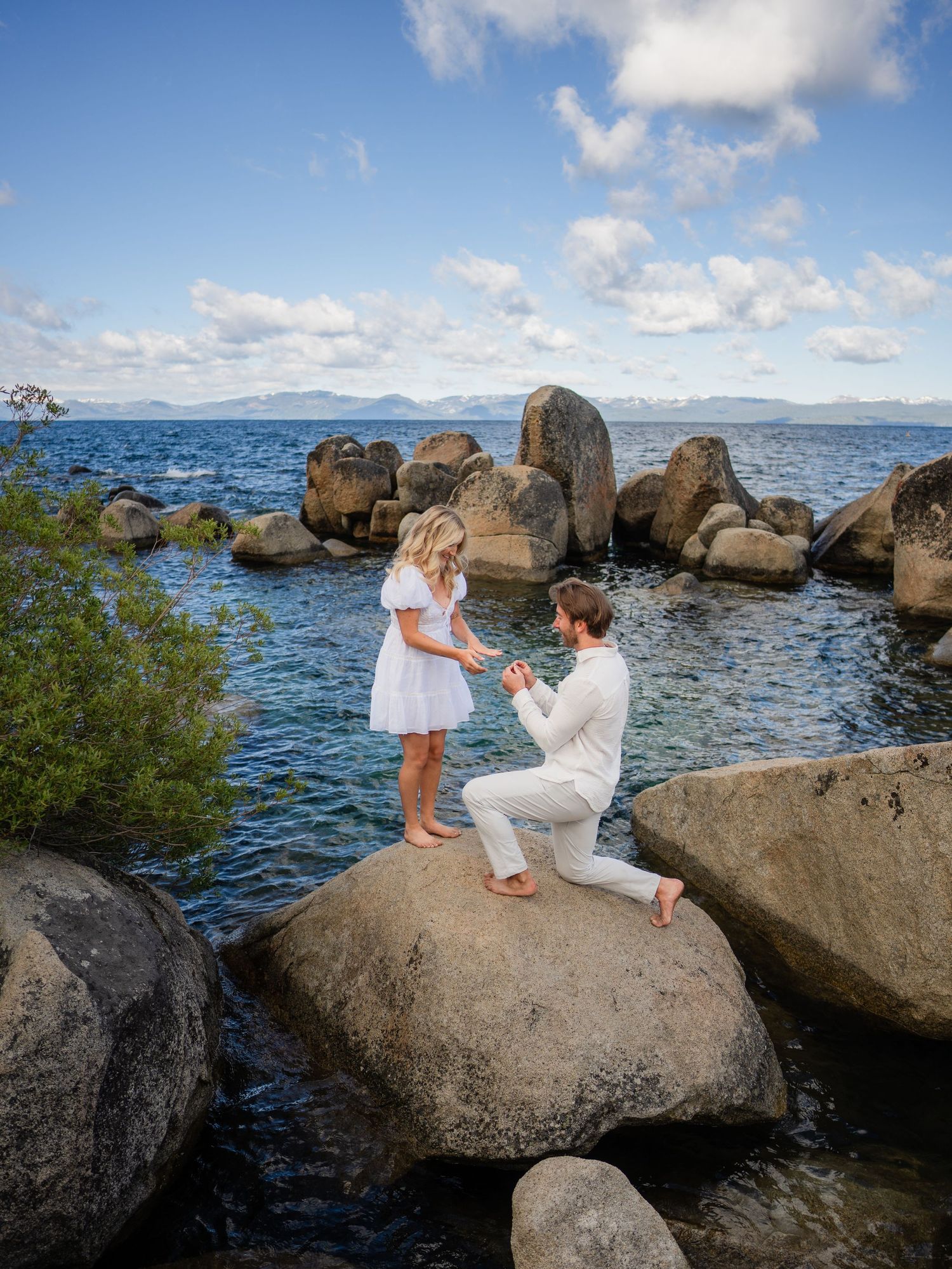A couple shares an intimate proposal moment on large rocks by Lake Tahoe with mountains visible in the background.