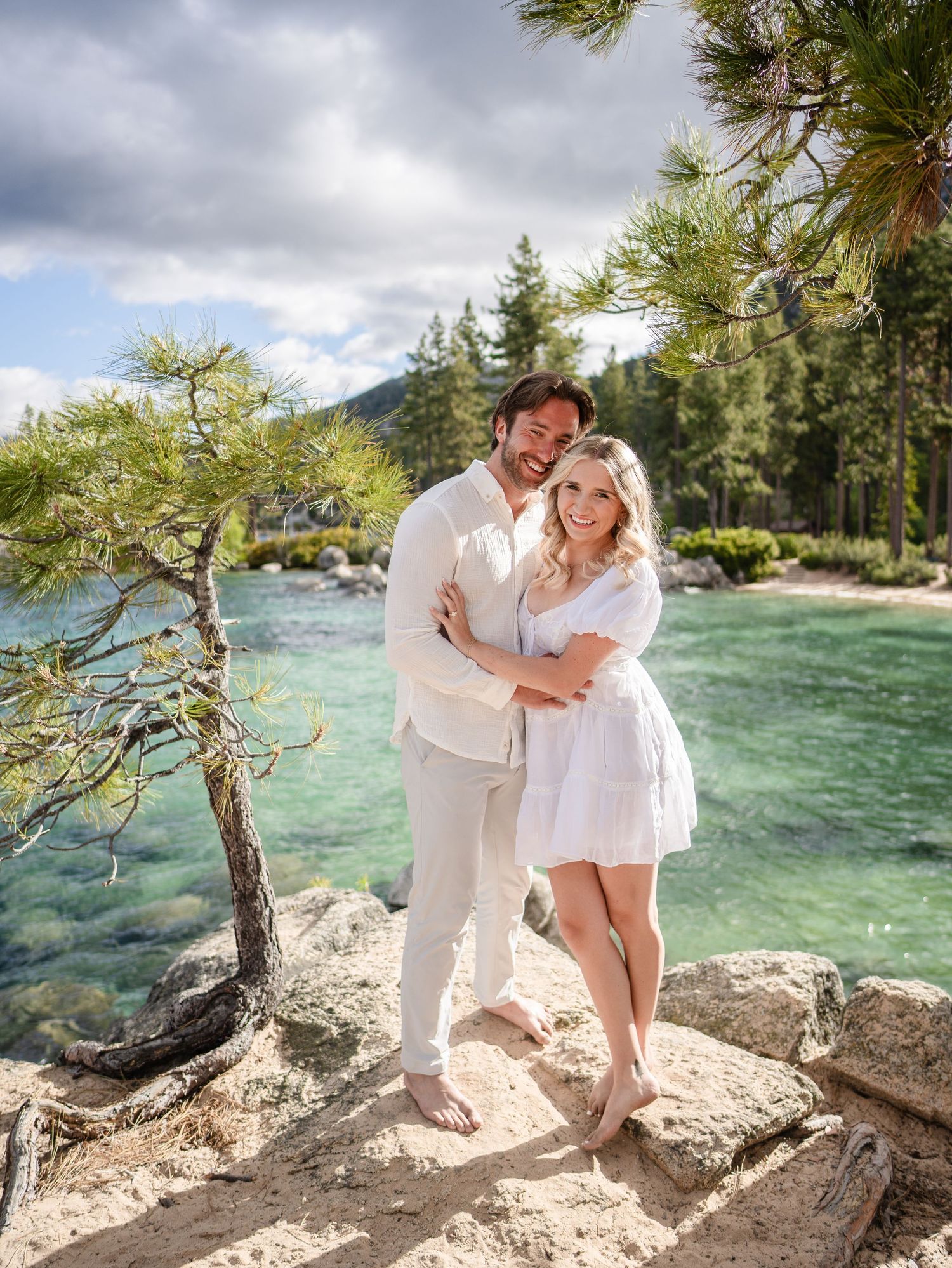 A couple in light summer outfits embraces on a rocky outcrop overlooking turquoise waters at Lake Tahoe.