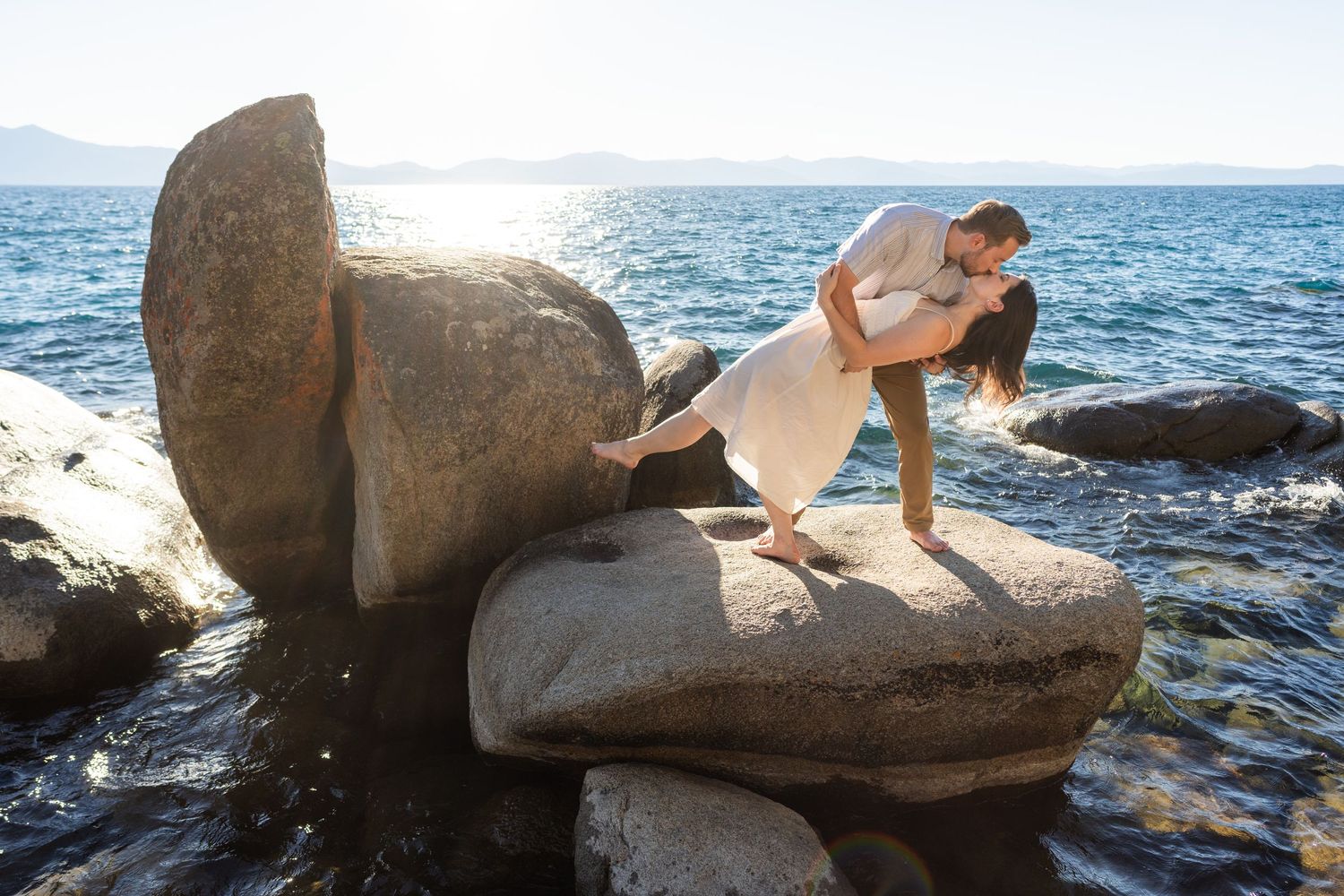 Two people sharing a romantic moment in Lake Tahoe on coastal rocks with crashing waves and bright sunlight in the background.