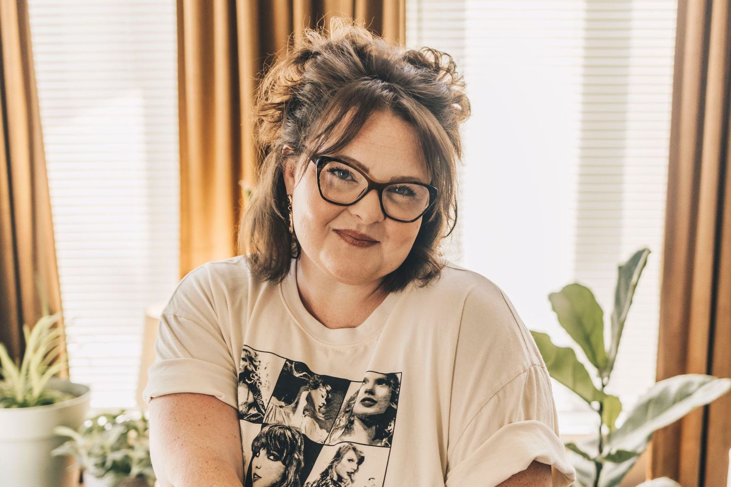 Smiling woman in Taylor Swift graphic tee posing among indoor plants near window.