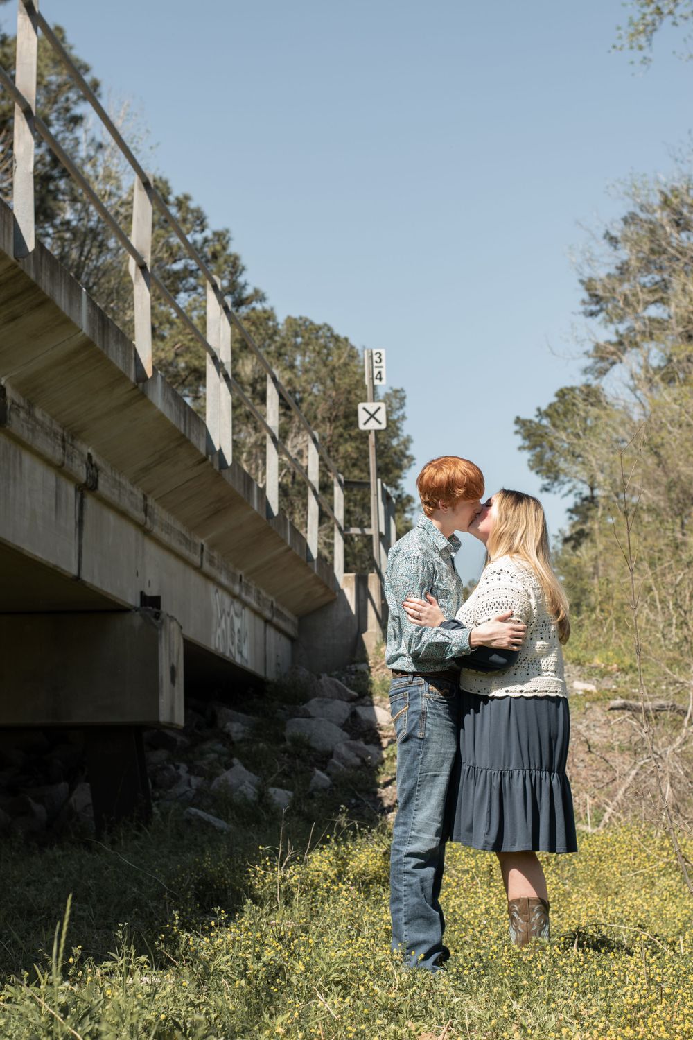A couple embraces near a rustic wooden bridge on a sunny spring day in a natural outdoor setting.