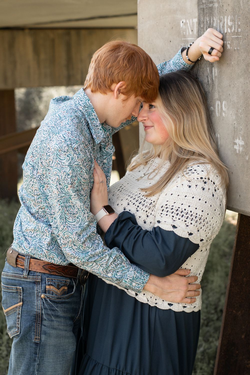 A romantic couple in casual attire share an intimate moment against a concrete wall.