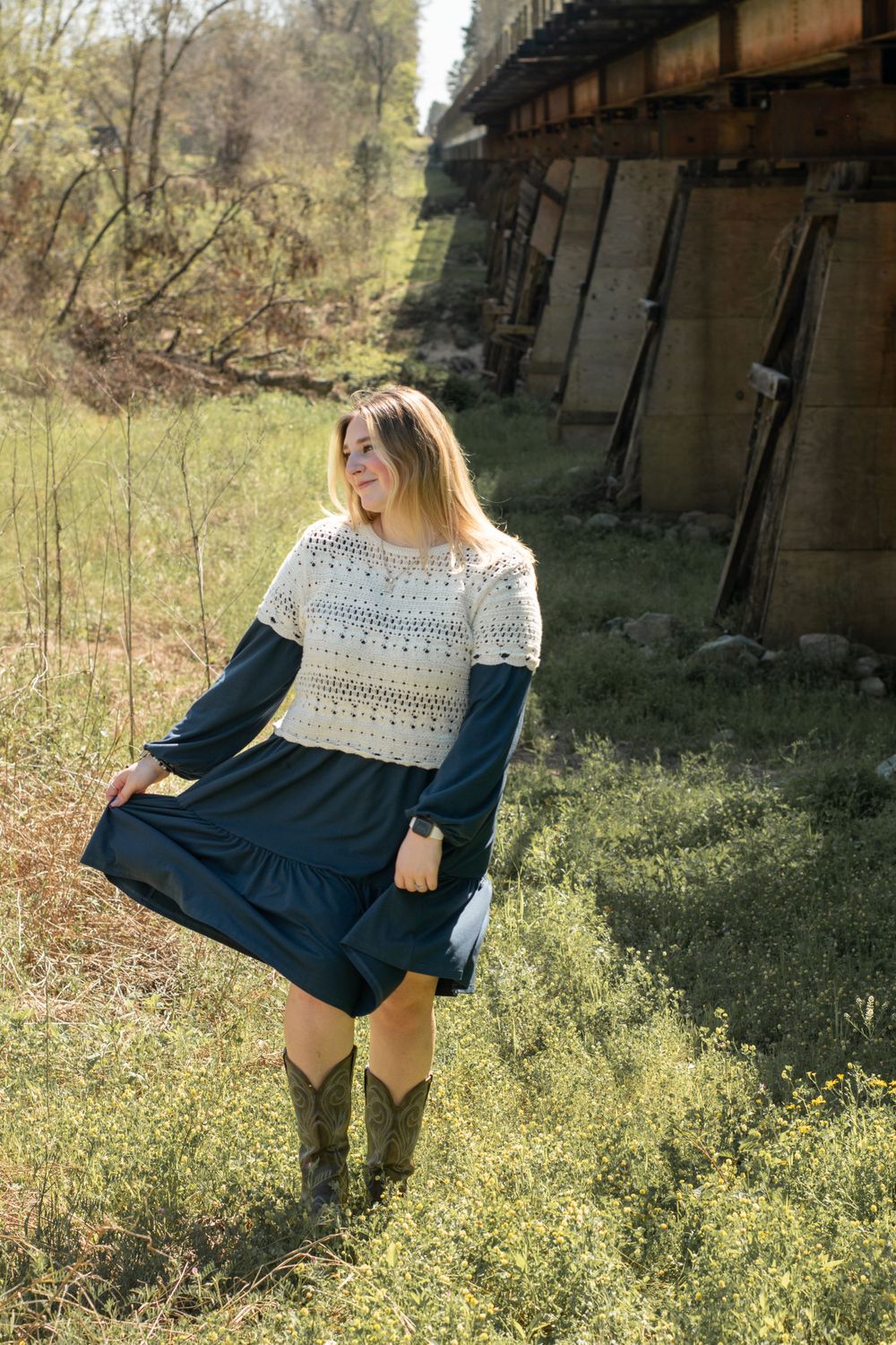 A person in brown boots and a navy dress with crochet detail poses in a rustic outdoor setting near a wooden structure.