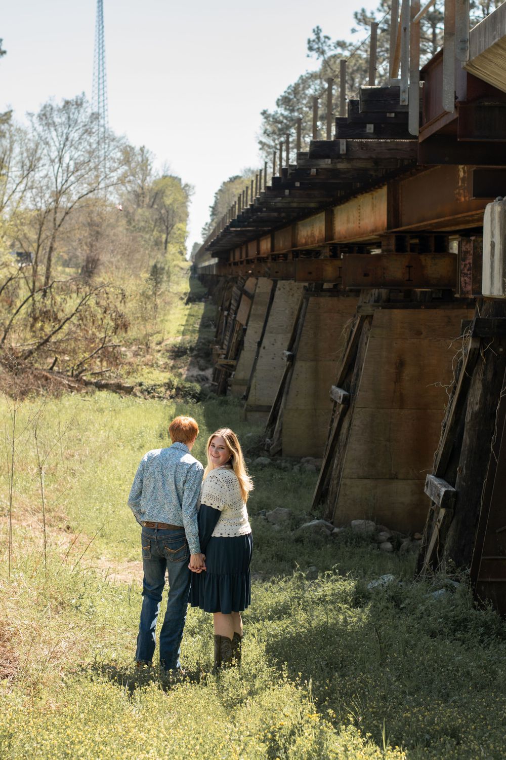 Two people stand together on grassy terrain beside an old wooden railway trestle bridge structure in a rustic outdoor setting.
