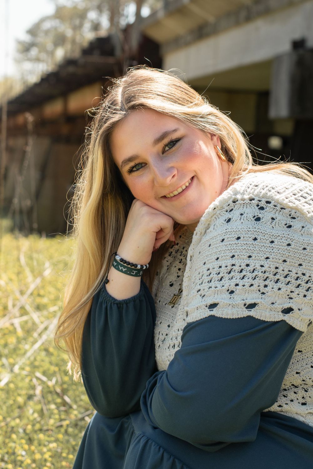 Smiling person with long blonde hair wearing a navy dress and white knit shawl poses outdoors on a sunny day.