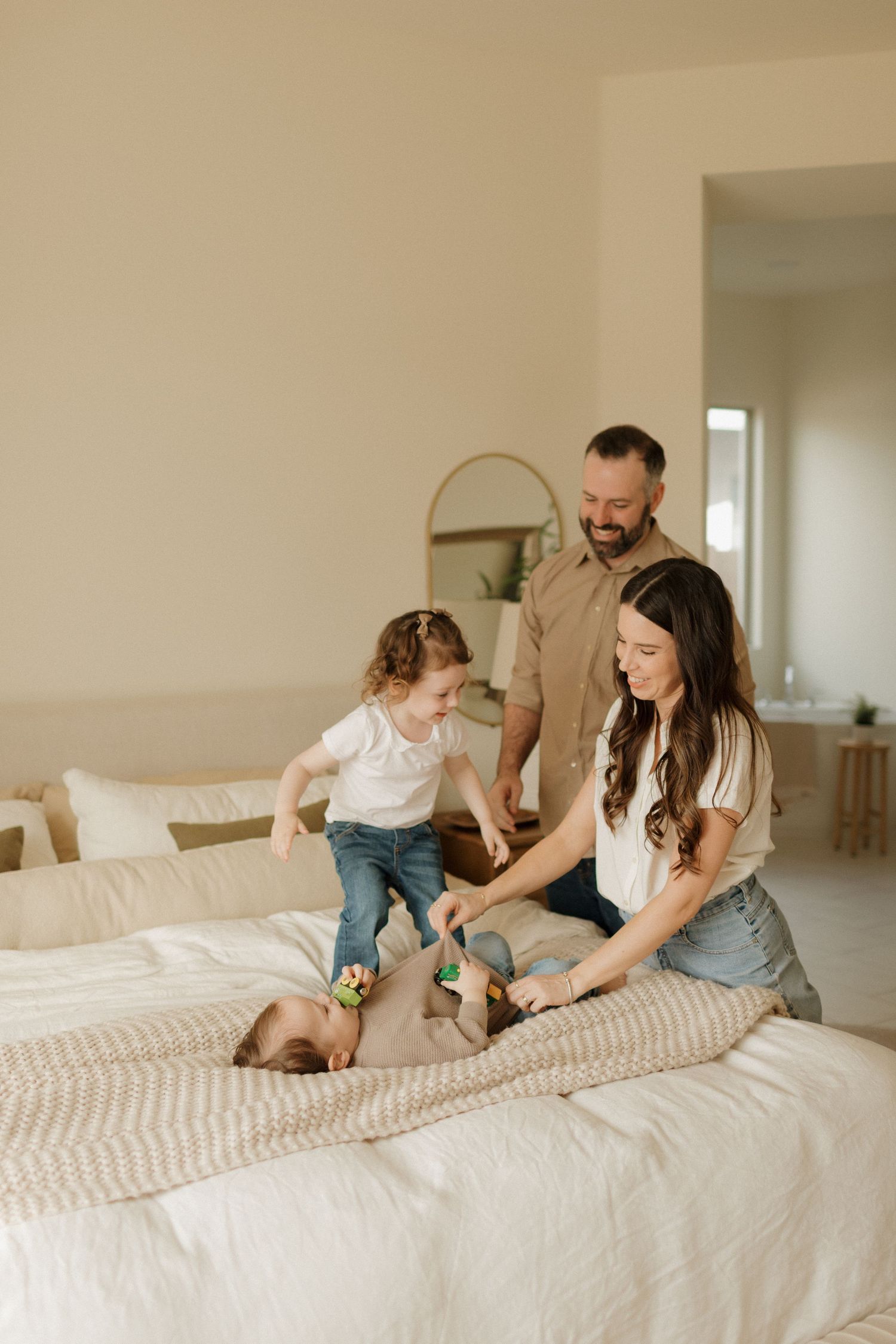 A joyful family moment playing together on a cozy bed with white linens in a minimal bedroom setting.