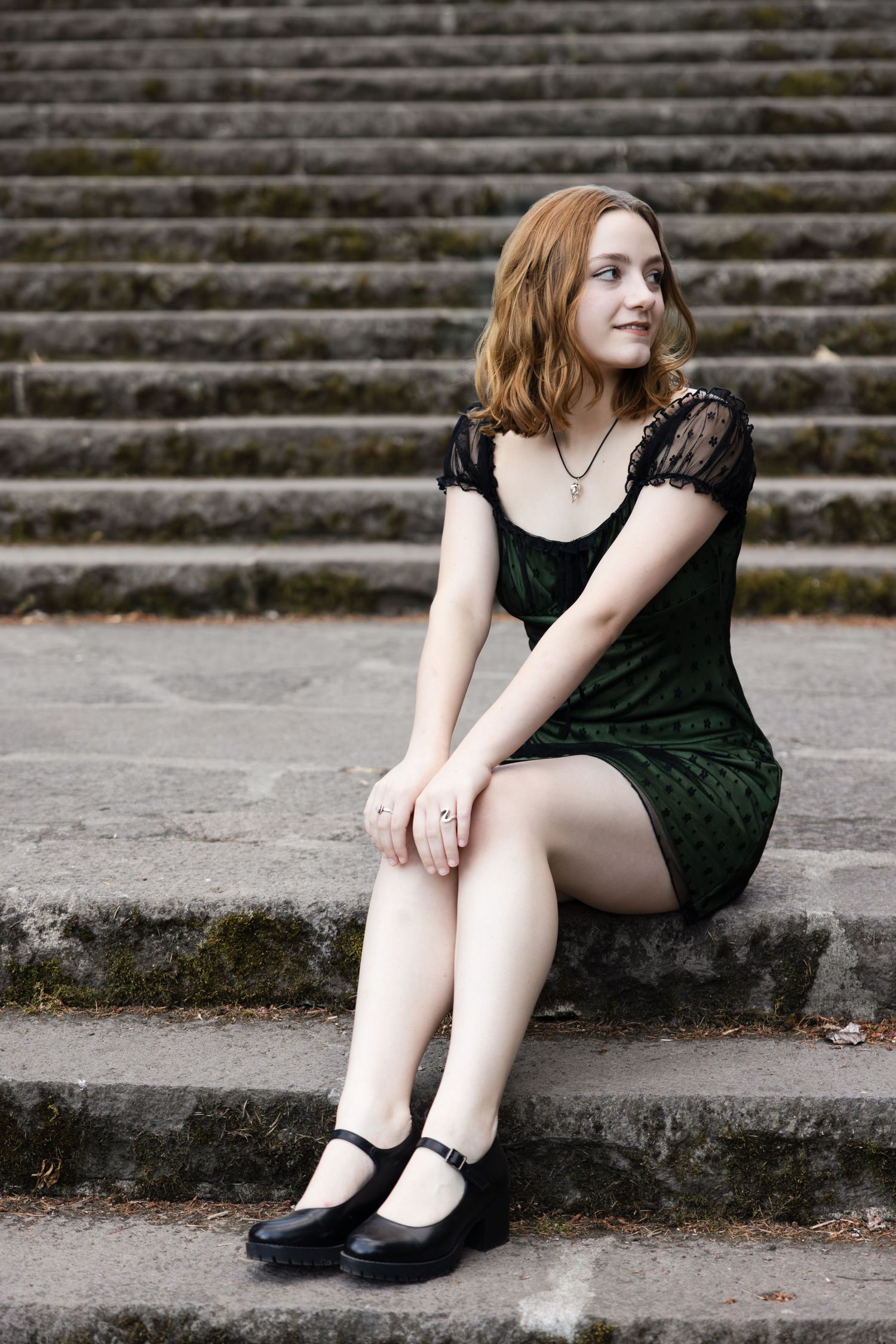 A person in a dark green dress sits on stone steps wearing black Mary Jane shoes.