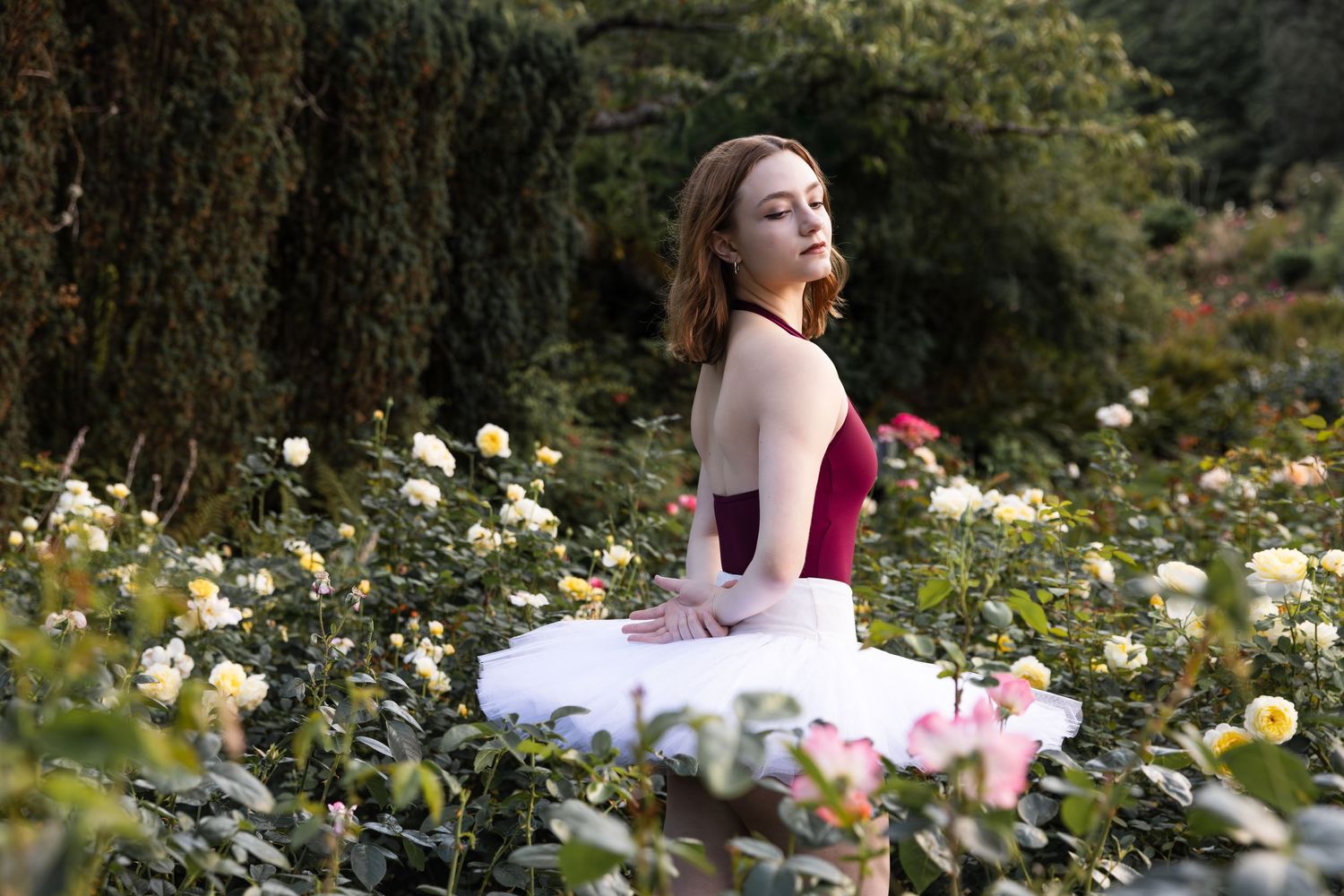 A ballet dancer in a pink tutu sits gracefully among yellow roses in a lush garden setting.
