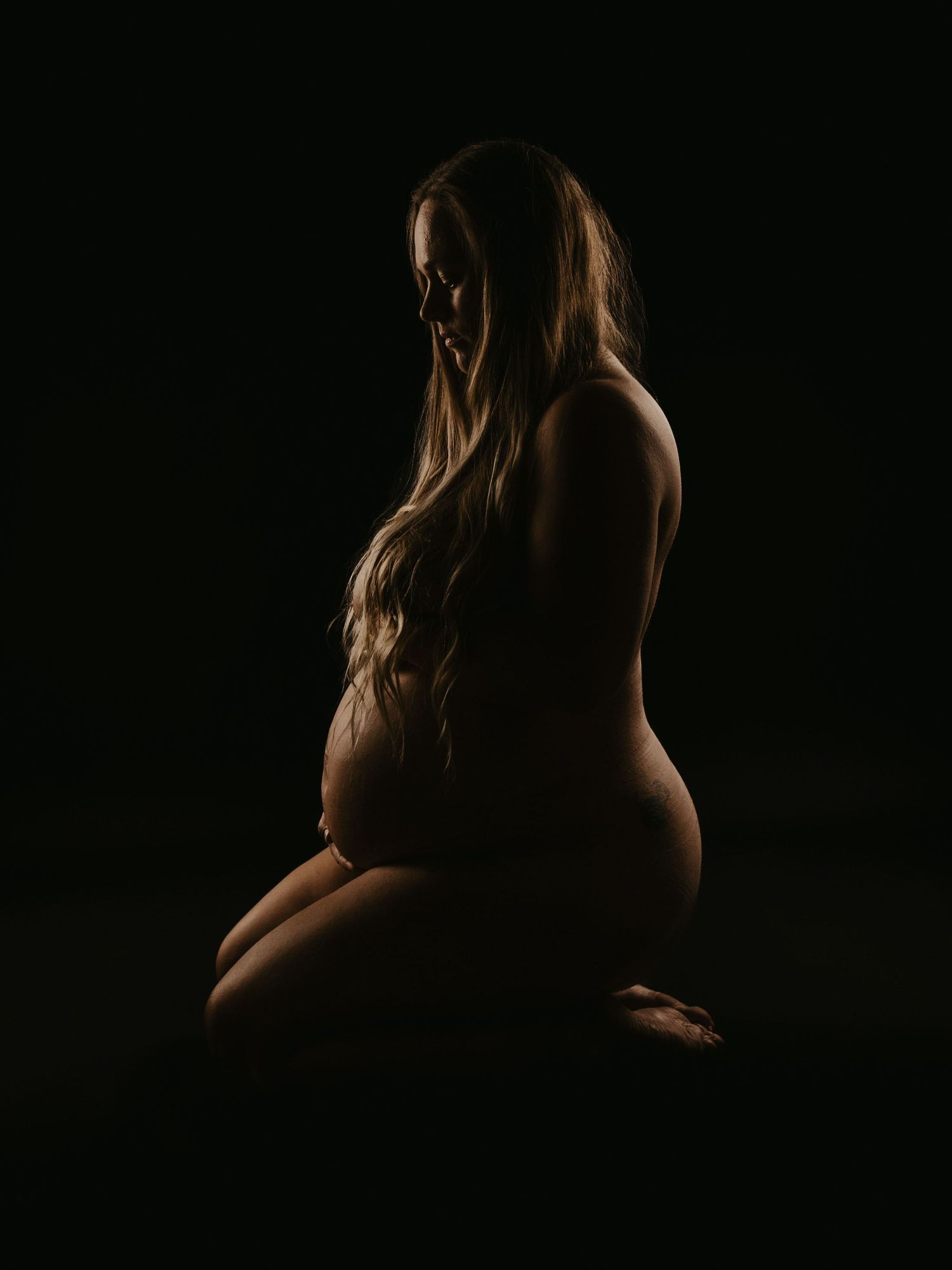 A dramatic silhouette maternity photo shows an expectant mother kneeling in a dark studio with rim lighting.