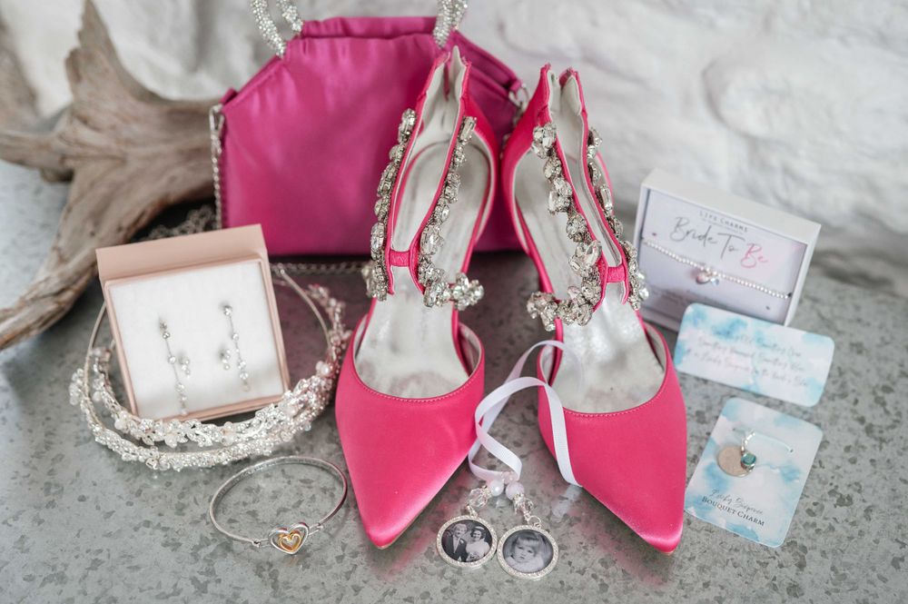 Pink bridal accessories including pointed heels, jewelry, and perfume boxes arranged on light fabric background.